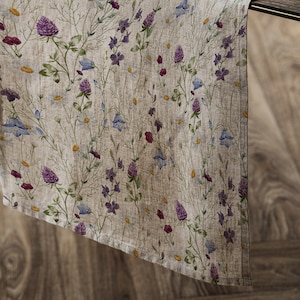 Puede incluir: Un camino de mesa de lino beige con un patrón floral con flores silvestres moradas, azules y verdes. El camino de mesa está colgando sobre el borde de una mesa de madera.