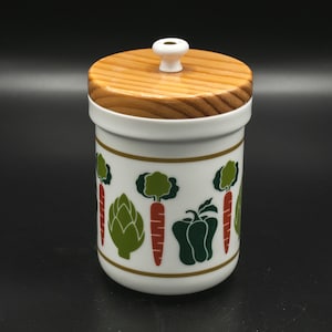 May include: White ceramic canister with a wooden lid and a green, red, and brown vegetable design. The canister has a gold trim around the top and bottom.
