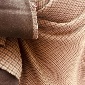 May include: Close-up of a brown and tan plaid fabric with a dark brown trim. The fabric has a classic pattern and a soft texture, with visible folds and creases. The trim appears to be a suede-like material.