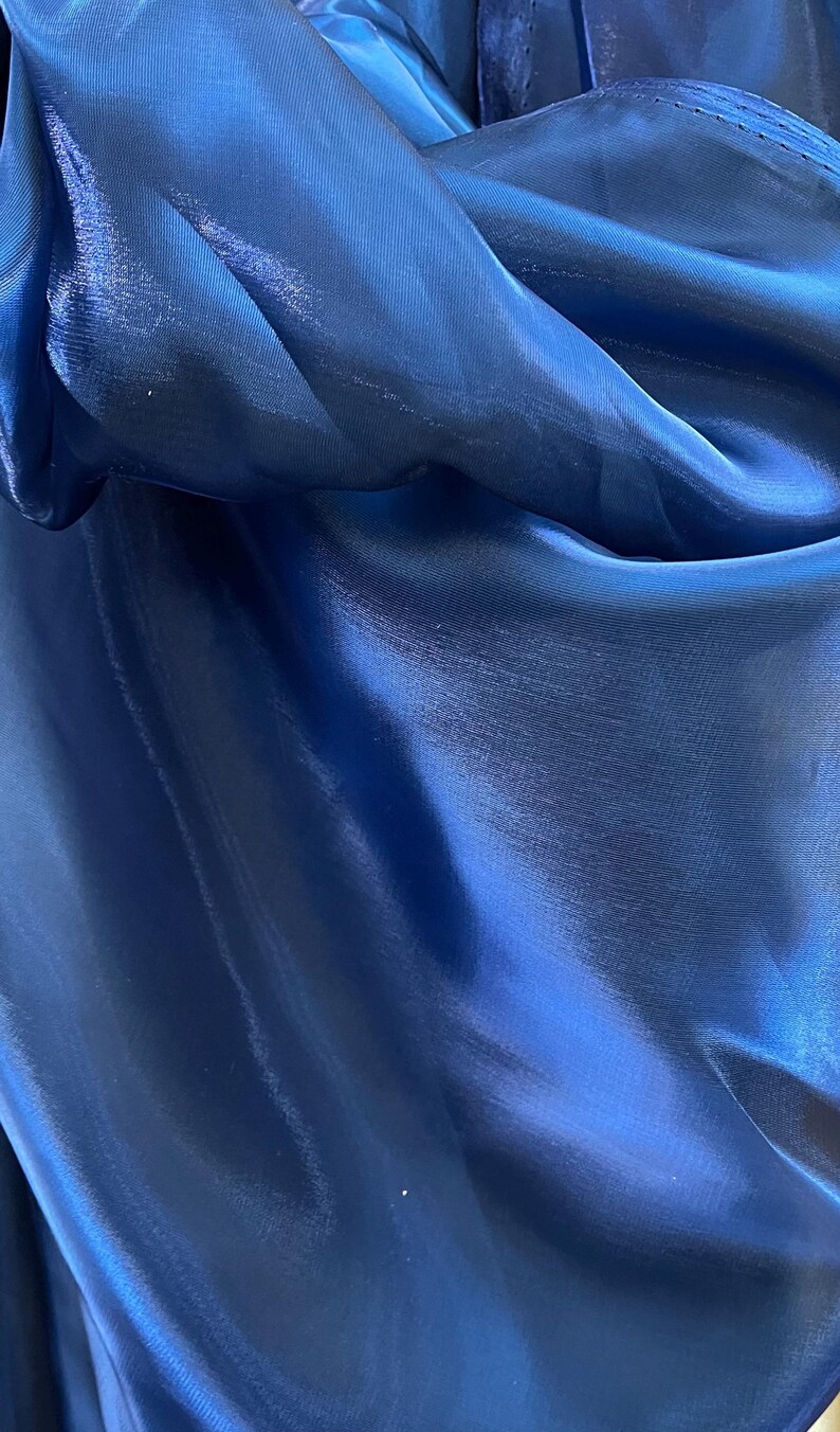 May include: Close-up of a deep blue fabric with a shiny, silky texture. The material is gathered and folded, creating shadows and highlights that emphasize its smooth surface. The fabric appears to be a flowing material.