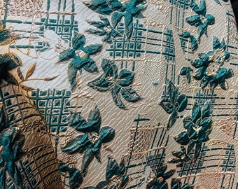 May include: Close-up of a textured fabric with a floral pattern. The fabric features teal-coloured raised floral designs and gold accents against a cream-coloured background. The design includes grid-like patterns and stylized floral elements.
