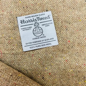 May include: Close-up of Harris Tweed fabric, a hand-woven cloth from the Outer Hebrides of Scotland. The fabric is a speckled tan color with flecks of red, yellow, and black. A label indicates it is 100% pure new wool.