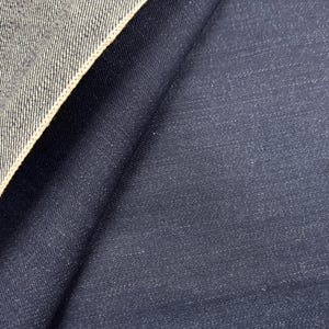 Puede incluir: Primer plano de tela vaquera. La imagen muestra un material de mezclilla azul oscuro con un borde de orillo de color más claro. La tela está doblada, revelando la textura y el tejido de la mezclilla.