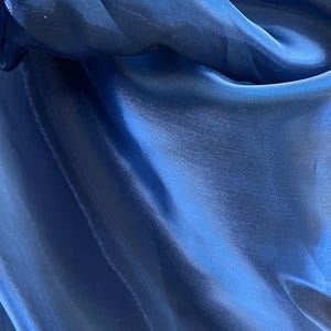 May include: Close-up of a flowing, deep blue fabric with a lustrous sheen. The material is draped, creating soft folds and highlights. The fabric's texture appears smooth and lightweight, reflecting light.