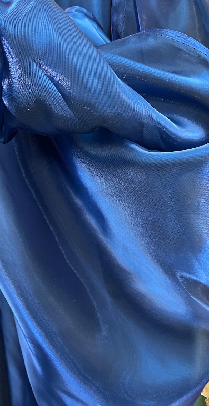 May include: Close-up of a flowing, deep blue fabric with a lustrous sheen. The material is draped, creating soft folds and highlights. The fabric's texture appears smooth and lightweight, reflecting light.