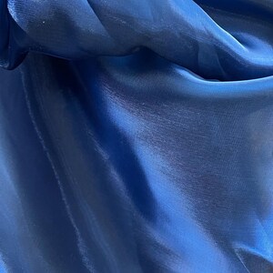 May include: Close-up of a deep blue fabric with a shiny, silky texture. The material is folded and draped, creating shadows and highlights that emphasize its smooth surface. The fabric appears to be a lightweight material, possibly for clothing or decorative purposes.