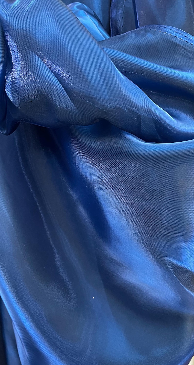 May include: Close-up of a deep blue fabric with a shiny, silky texture. The material is folded and draped, creating shadows and highlights that emphasize its smooth surface. The fabric appears to be a lightweight material, possibly for clothing or decorative purposes.