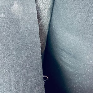 Può includere: Primo piano di un tessuto nero con una texture sottile. Il tessuto è piegato a metà, rivelando una cucitura lungo la piega.