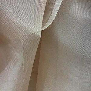 Può includere: Primo piano di un tessuto beige trasparente con un fine motivo a rete. Il tessuto è piegato a metà, creando una piega al centro. I bordi del tessuto sono cuciti con filo bianco.