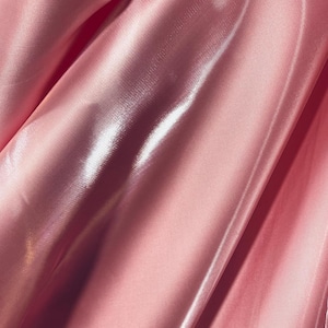 Peut inclure: Gros plan sur un tissu satiné rose avec un éclat brillant. Le tissu est drapé, créant des plis et des reflets doux. La couleur est un rose clair et rosé, et la texture semble lisse et luxueuse.