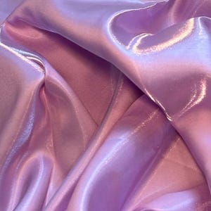 Peut inclure: Gros plan sur un tissu fluide dans des tons lavande et rose. La matière a une texture brillante, semblable à du satin, avec des plis doux et des reflets qui créent une sensation de profondeur et de mouvement. L'effet global est lisse et élégant.