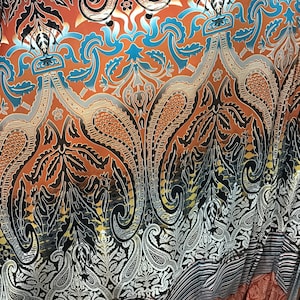 May include: A fabric with a detailed paisley and floral pattern. The design features shades of orange, black, blue, and silver. The fabric has a striped border at the bottom and is likely used for clothing or home decor.