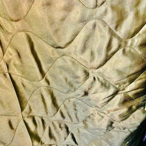 May include: Close-up of olive green quilted fabric. The fabric has a wavy, stitched pattern across its surface. The material appears smooth and slightly shiny, reflecting light. The overall effect is one of texture and depth.