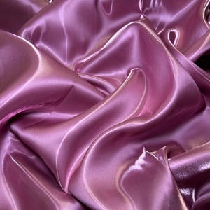 Peut inclure: Gros plan sur un tissu fluide de couleur rose pâle. Le matériau présente un aspect brillant, semblable à du satin, avec des plis et des courbes qui créent des reflets et des ombres. Le tissu semble lisse et délicat, idéal pour l'habillement ou la décoration intérieure.
