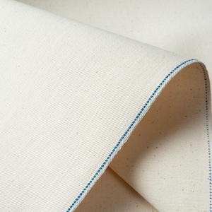 Puede incluir: Primer plano de una tela de lona blanquecina doblada, con un borde cosido azul. La tela tiene un sutil patrón de tejido diagonal y una superficie ligeramente texturizada. El borde está cuidadosamente terminado con una fina línea de costura azul.