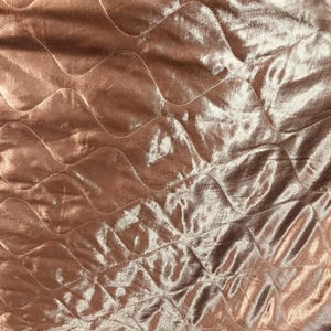 Puede incluir: Primer plano de una tela acolchada en un cálido color oro rosa. La tela tiene un brillo suave y lustroso y está cosida con un patrón de diamantes ondulados. La textura sugiere un material afelpado, posiblemente terciopelo o satén.
