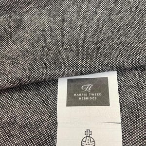 May include: Close-up of a textured fabric in black and white. The fabric has a woven appearance with a speckled pattern. A tag with the text "HARRIS TWEED HEBRIDES" and a logo is visible.