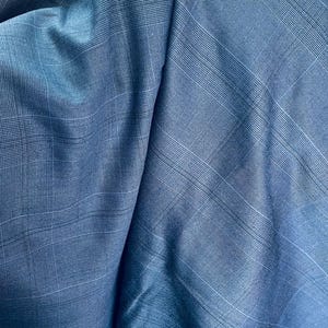 May include: Close-up of a blue fabric with a plaid pattern. The fabric has a subtle sheen and is folded, creating shadows and highlights. The weave is visible, adding texture to the material. The overall tone is a cool, sophisticated blue.