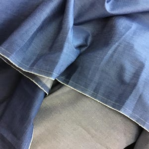 May include: Close-up of folded blue fabric with white stitching along the edges. The fabric has a textured appearance and a slight sheen. The folds create shadows and variations in the color, highlighting the material's depth.