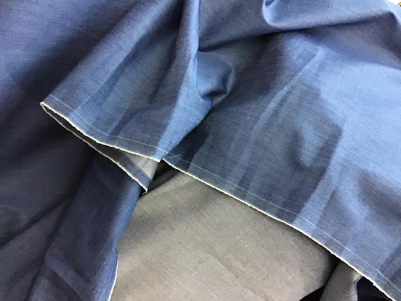 May include: Close-up of folded blue fabric with white stitching along the edges. The fabric has a textured appearance and a slight sheen. The folds create shadows and variations in the color, highlighting the material's depth.