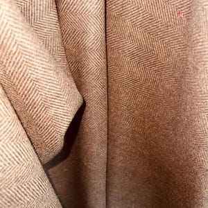 May include: Close-up of a brown fabric with a herringbone pattern. The fabric is folded, creating shadows and depth. The weave is visible, showing the texture and detail of the material. The color is a warm, earthy tone.