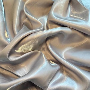 Peut inclure: Gros plan sur un tissu argenté clair et chatoyant avec un aspect satiné. Le tissu est drapé en plis souples, créant des reflets et des ombres. Le matériau semble léger et fluide, adapté à l'habillement ou à la décoration.