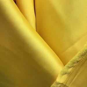 Puede incluir: Primer plano de una tela de color amarillo brillante con una textura suave y sedosa. La tela está doblada de forma que crea una V.