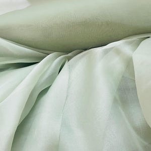 Pode incluir: Close-up de um tecido verde claro com uma aparência macia e translúcida. O tecido é drapeado em dobras suaves, criando um efeito em camadas. O material tem um brilho sutil, sugerindo uma textura delicada.