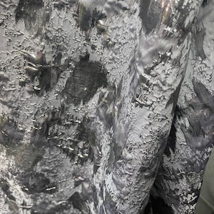 May include: Close-up of a textured fabric with a metallic sheen. The fabric is primarily gray with darker, shadowy areas and highlights of silver. The surface has a raised, irregular pattern, creating a tactile appearance.