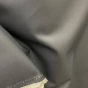 May include: A close-up shot of a dark gray, smooth fabric. The material is folded, creating shadows and highlights that emphasize its texture. The edge of the fabric reveals a white lining.