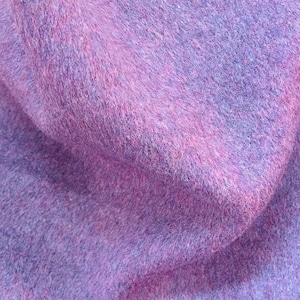 May include: A piece of felt fabric featuring a mottled blend of purple, lavender, and magenta fibers. The material has a dense, fuzzy, and non-woven texture with visible variations in color density throughout the surface of the textile.