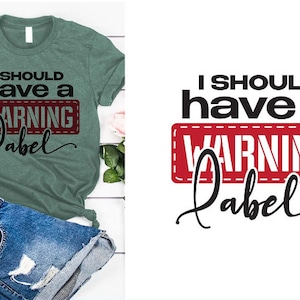 Könnte beinhalten: Ein grünes T-Shirt mit dem Text "I should have a warning label" in schwarzer Schrift mit einem roten Warnhinweis mit weißen Nähten.
