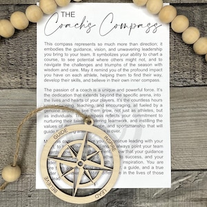 May include: Wooden compass ornament with the text "The Coach's Compass" and the words "Guide", "Inspire", "Lead", and "Believe". The compass is attached to a string and a wooden bead. A white card with text is behind the compass.