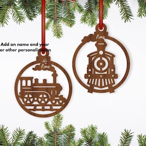May include: Two wooden train-themed Christmas ornaments hanging from red ribbons. Each ornament features a train design within a circular frame. The text "Name" is visible on the top of each ornament, suggesting personalization. The ornaments are displayed against a white background.