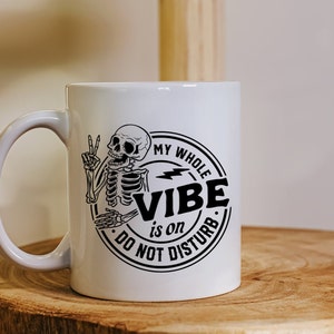 May include: White ceramic coffee mug with a black and white graphic of a skeleton with a peace sign and the text "My Whole Vibe is on Do Not Disturb".