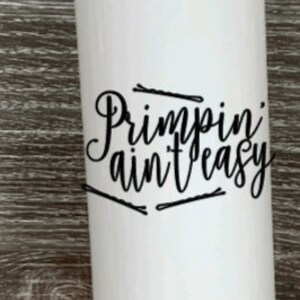 May include: White stainless steel tumbler with a black inscription that reads "Primpin' ain't easy" and a few black hair clips around the text.