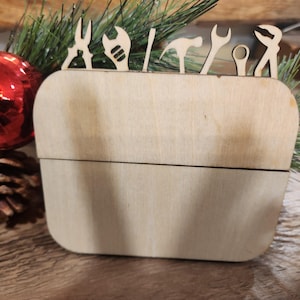 May include: A wooden box with a hinged lid. The lid has a cutout design of various tools, including a wrench, hammer, screwdriver, and pliers. The box is made of light-colored wood and has a natural finish.