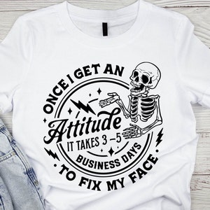 May include: White t-shirt with a black circle graphic featuring a skeleton with the text "Once I get an Attitude it takes 3-5 business days to fix my face."