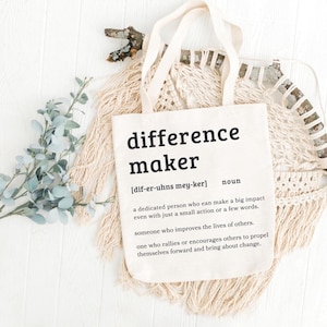 May include: A cream-colored tote bag with the word "difference maker" printed in black. The definition of "difference maker" is printed below the word. The bag is on a white surface with decorative elements.