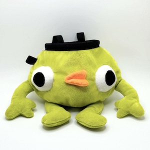 Może przedstawiać: Limonkowo-zielony pluszowy worek na magnezję w kształcie kreskówkowej postaci z dużymi białymi i czarnymi oczami, pomarańczowym dziobem oraz małymi zielonymi ramionami i stopami. Worek ma czarny otwór i dwie czarne pętle.