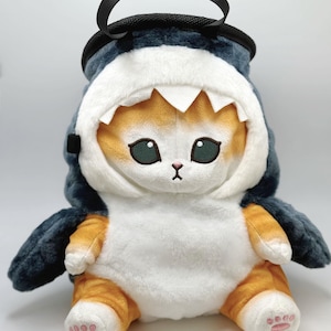 May include: A plush toy cat wearing a blue shark costume with a black handle on top. The cat has orange and white fur and pink paw pads.