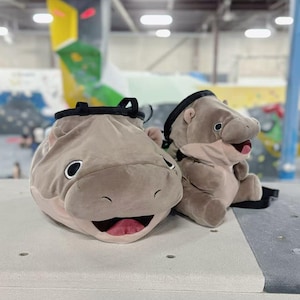 May include: Two plush climbing chalk bags shaped like hippos. One is lying down, the other is sitting up. Both are light brown with black accents and open mouths. The bags have a drawstring closure at the top.