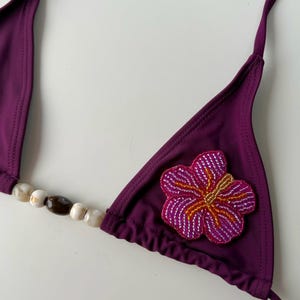 Può includere: Un top bikini color prugna con una stringa di perline e un'applicazione floreale. Il top a triangolo presenta una stringa di perline con perline bianche e marroni. Un'applicazione floreale rosa, arancione e bianca è cucita sulla coppa destra.
