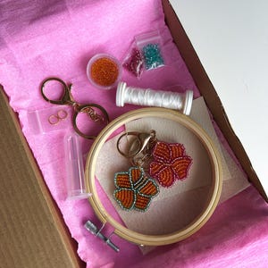 May include: A craft kit containing keychains, beads, thread, and an embroidery hoop. The keychains feature beaded flower designs in orange and red. The kit includes small bags of beads in various colours and a spool of white thread.