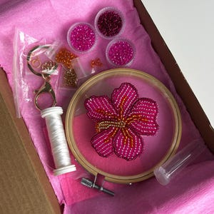 May include: A craft kit containing a pink flower embroidery hoop, beads, thread, and a keychain. The flower is made with pink and gold beads. The kit includes small containers of pink and red beads, all inside a pink box.