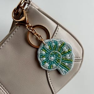 May include: A beaded seashell keychain with a gold-colored clasp and chain. The seashell design features teal, green, and white beads on a silver-colored backing. The keychain is attached to a light beige purse.