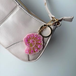 May include: A pink and gold beaded seashell keychain attached to a light gray purse. The keychain features a detailed design with pink beads outlining the shell shape and gold beads filling the center. The purse has a gold zipper and strap.
