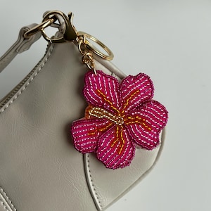 Puede incluir: Un llavero de flor rosa con cuentas y detalles dorados. La flor tiene un diseño detallado con costuras blancas y amarillas. El llavero está sujeto a un bolso gris claro con un cierre dorado.