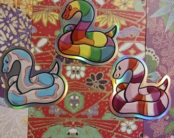 May include: Three iridescent vinyl stickers featuring different colored snakes. The first snake is rainbow colored, the second is blue and white, and the third is pink and red.