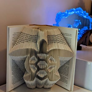 May include: A book with pages folded into the shape of a flower. The flower is centered on the open book, with the pages forming the petals and center. The book is on a white shelf.